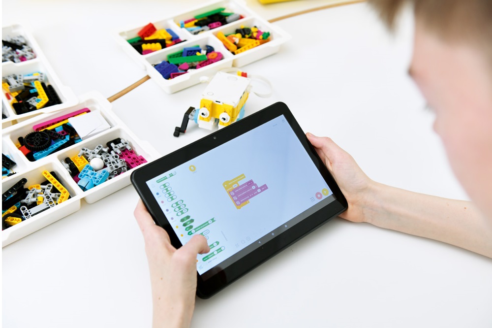 Chłopiec programuje zestaw lego education spike prime w aplikacji lego na tablecie, na białym stole klocki lego i model robota